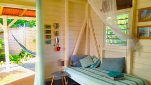 A room with a bed, a small table, a chair, a hammock outside, and a window with a view of greenery.