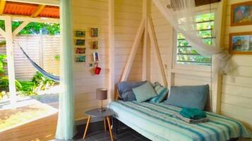 A room with a bed, a small table, a chair, a hammock outside, and a window with a view of greenery.