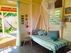 A room with a bed, a small table, a chair, a hammock outside, and a window with a view of greenery.