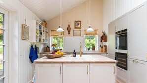 Une cuisine moderne avec des meubles blancs, un îlot central, des luminaires suspendus et une grande fenêtre qui laisse entrer la lumière naturelle.