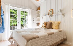 Ein Schlafzimmer mit einem großen Bett, einem Fenster mit Vorhängen, einem Bücherregal und einem Kleiderhaken.