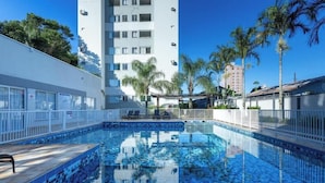 A swimming pool with a tiled deck, surrounded by a white fence, palm trees, and a high-rise building in the background. - Apt With Ocean View in Cond With Pool Ori1001 (Picarras)