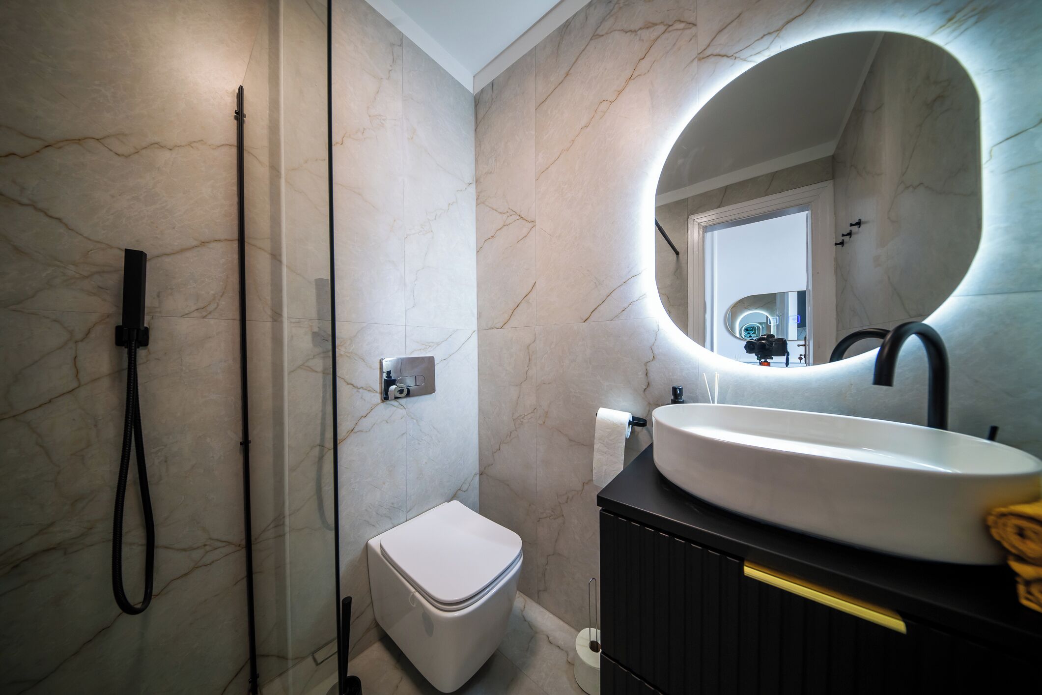 Un baño moderno con pared de mármol, espejo redondo, inodoro blanco y tocador negro con lavabo.