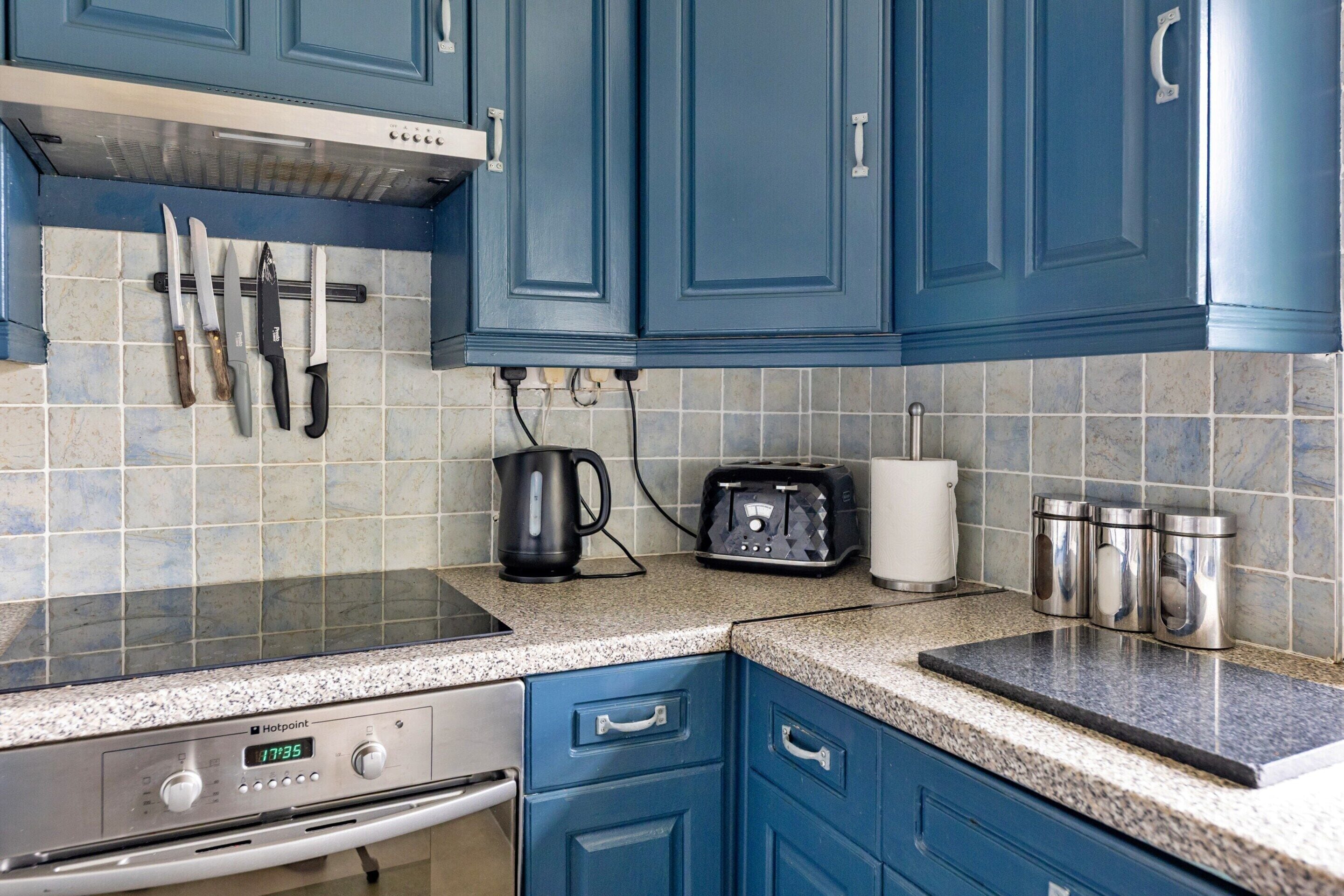 A kitchen with blue cabinets, a stove, a kettle, a toaster, and a trash can.