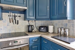 A kitchen with blue cabinets, a stove, a kettle, a toaster, and a trash can.