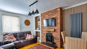 A living room with a brick fireplace, a leather sofa, a television mounted on the wall, and a patterned rug. - Holiday Cottage With Conwy Castle View (Conwy)