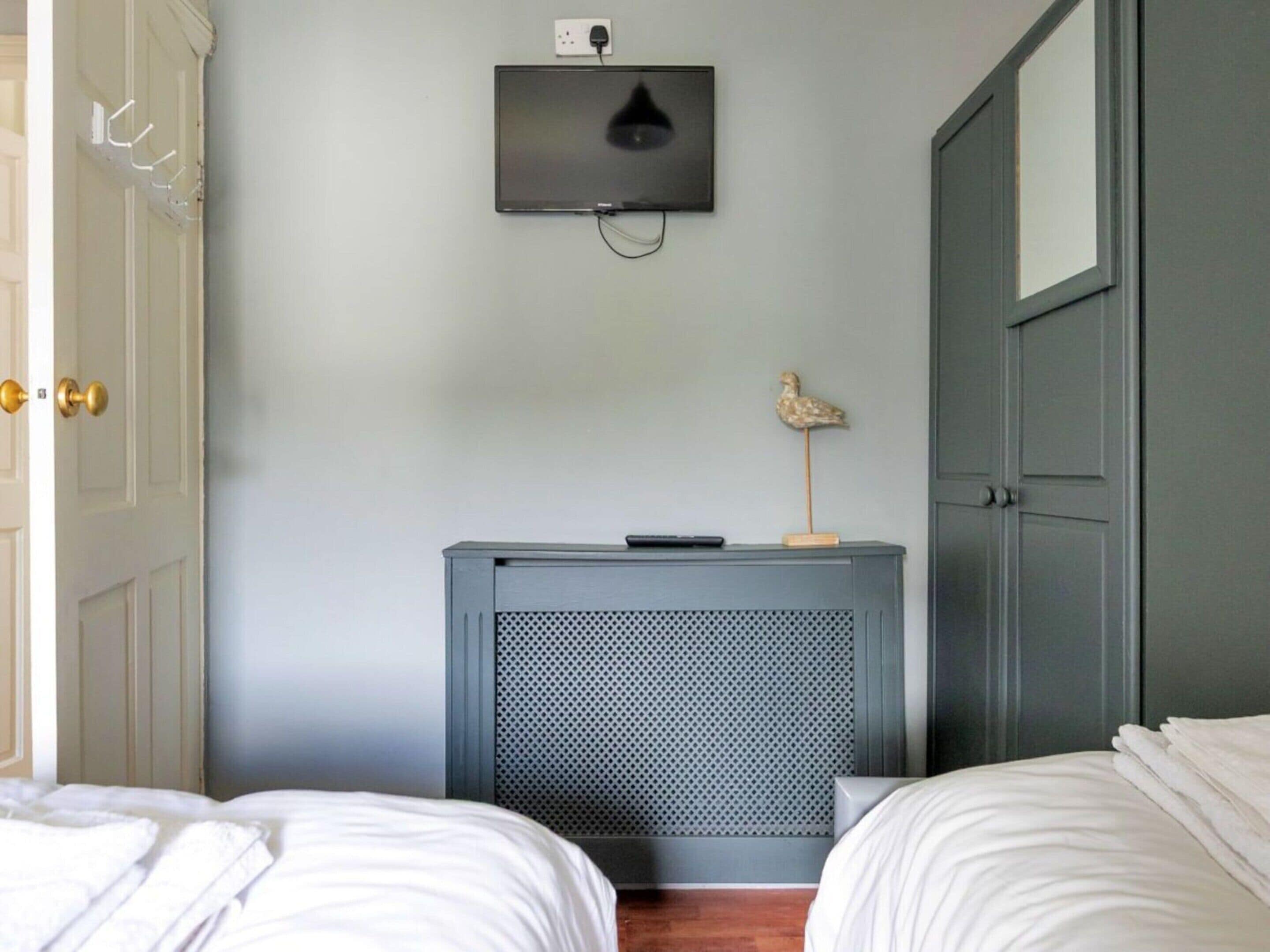 Quarto com duas camas, televisão na parede, uma mesinha com um objeto decorativo e uma porta com puxador dourado.