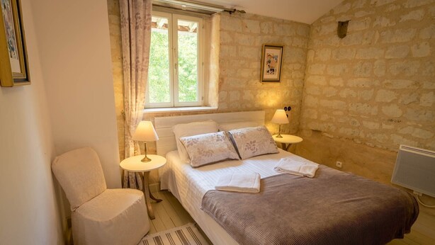 Une chambre à coucher avec un lit, deux lampes de chevet, une chaise, une fenêtre avec des rideaux et un mur en pierre.