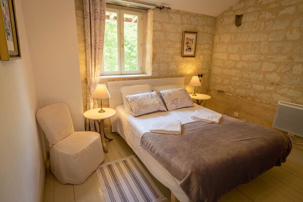 A bedroom with a bed, two bedside lamps, a chair, a window with curtains, and a stone wall.