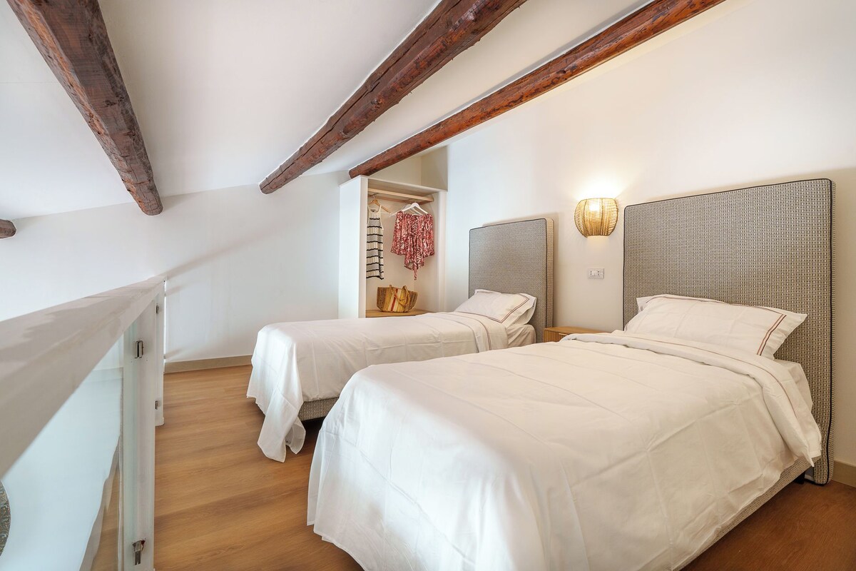 Een slaapkamer met twee bedden, houten balken in het plafond, een hanglamp en een kledingkast met kleding.