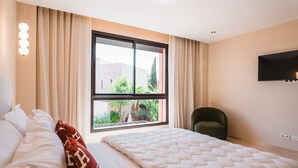 A hotel room with a bed, a chair, a large window with a view of greenery, and a mounted TV. - Riad Koutobia – Al Maaden Golf – Marrakech (Marrakech)