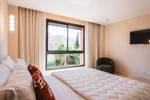 Ein Hotelzimmer mit Bett, Sessel, großem Fenster und Blick ins Grüne sowie einem wandmontierten Fernseher.