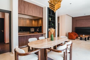 A modern living room with a dining area, featuring a wooden dining table, white cushioned chairs, a green vase with dried plants, and a unique hanging light fixture. - Riad Koutobia – Al Maaden Golf – Marrakech (Marrakech)