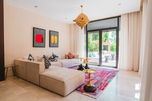 A modern living room with a beige sofa, patterned cushions, a glass coffee table, and a vibrant rug. - Riad Koutobia – Al Maaden Golf – Marrakech (Marrakech)