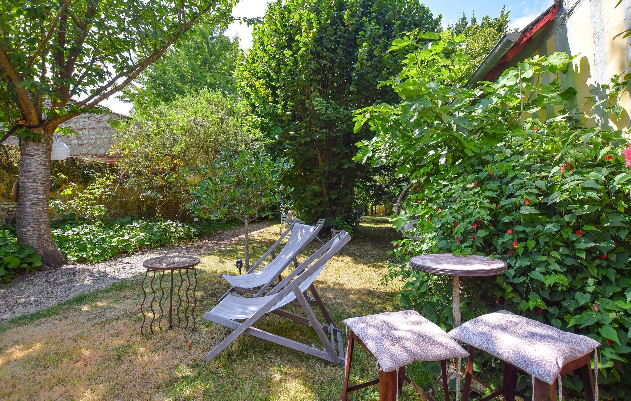 Un giardino con una poltrona da esterno in legno, due tavolini con cuscini decorati e diverse piante, tra cui alberi e arbusti.