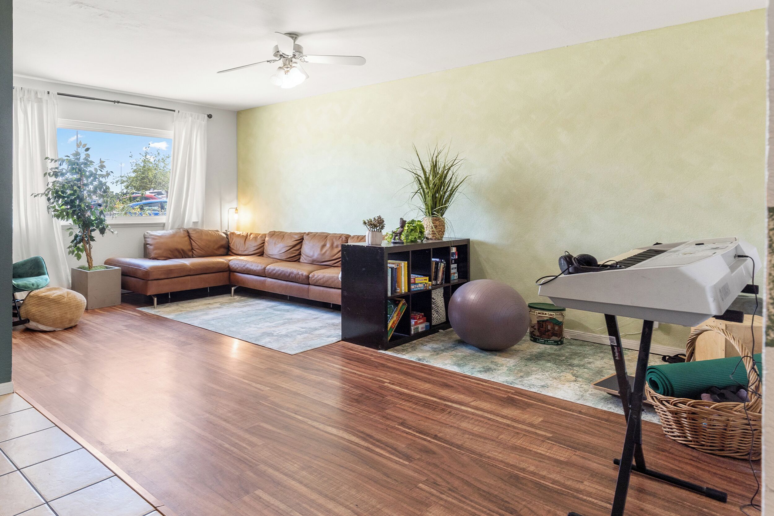 A living room with a brown leather sofa, a bookshelf, a plant, a yoga ball, and a piano.