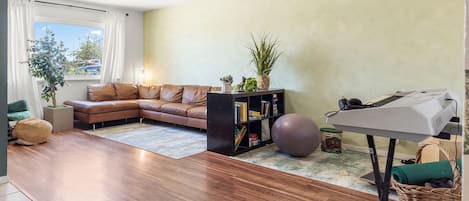 A living room with a brown leather sofa, a bookshelf, a plant, a yoga ball, and a piano.