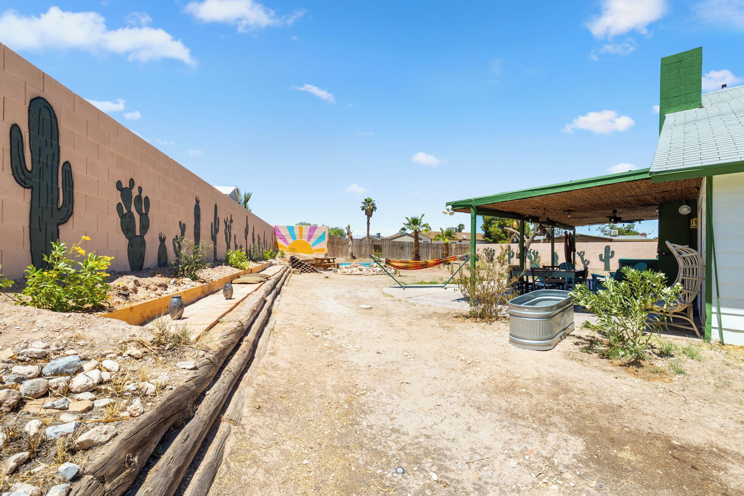 Un paysage désertique avec un bâtiment dont les murs sont décorés de motifs de cactus, une structure à toit de chaume et des sièges en plein air.