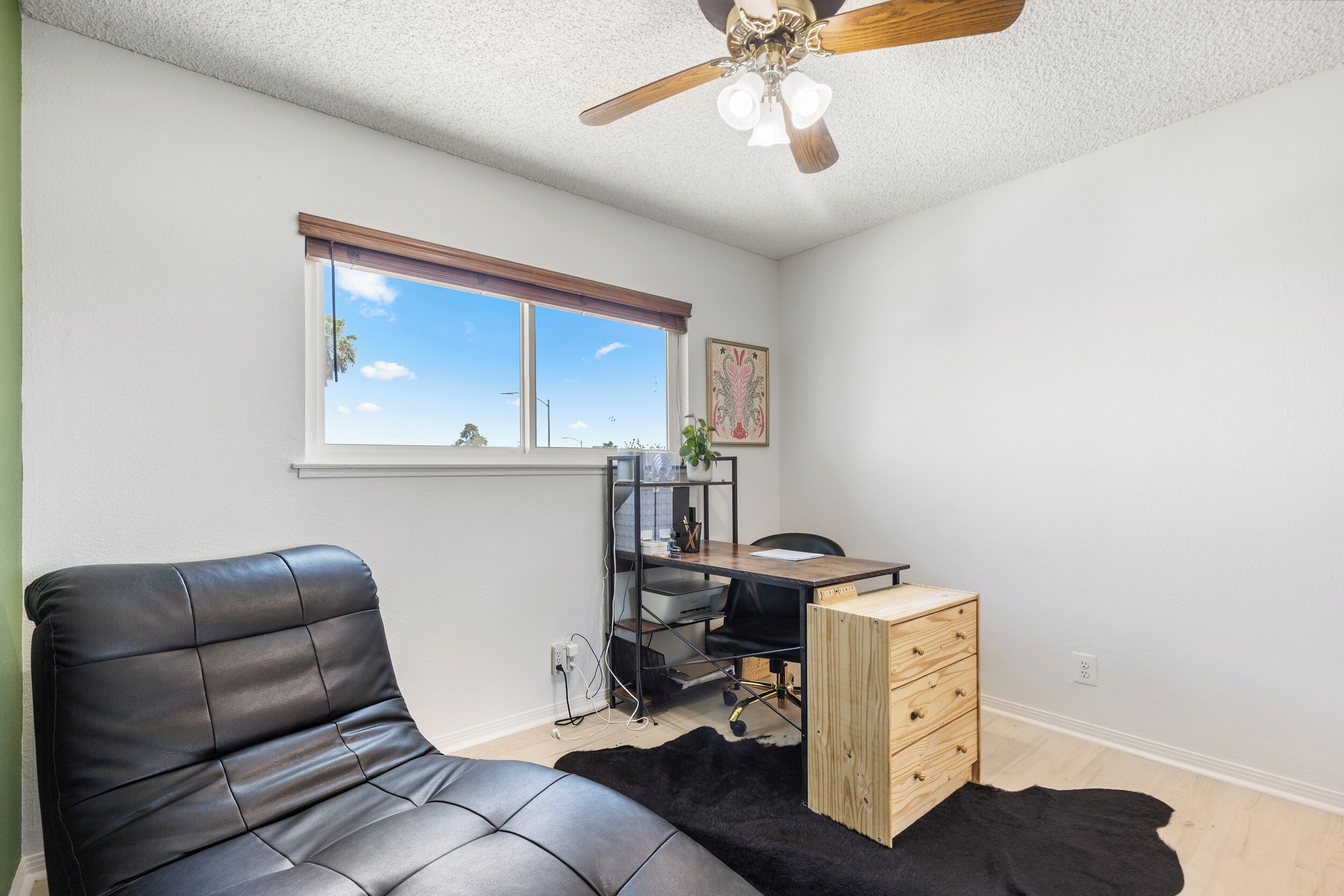 A room with a ceiling fan, a desk with a chair, a window with blinds, and a black leather sofa.