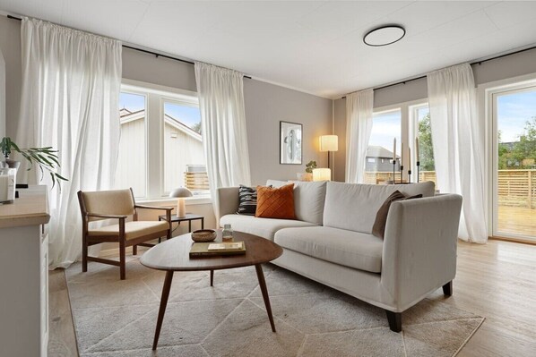 A living room with a sofa, a coffee table, and a chair. There is a window with curtains, a lamp, and a plant. - Exclusive South Tromsøya home peaceful & central (Tromsø)