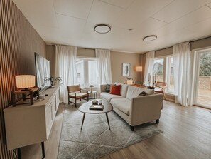 A modern living room with a sofa, coffee table, and a TV. There are curtains, a lamp, and a plant. - Exclusive South Tromsøya home peaceful & central (Tromsø)