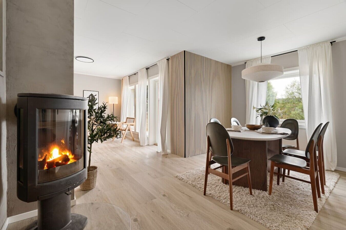 Un salon moderne doté d’une cheminée, d’un coin repas avec des chaises et une table, et d’une grande fenêtre avec des rideaux.