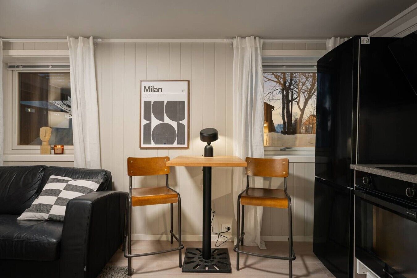 Un espace de vie compact comprenant un canapé en cuir noir, une table en bois avec deux chaises, une œuvre d’art encadrée accrochée au mur et un meuble TV noir.
