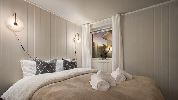 A neatly made bed with a beige blanket and pillows, a bedside table with a lamp, and a window with curtains. - Sydpol Designer Home Deluxe Apartment (Tromsø)