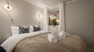 A neatly made bed with a beige blanket and pillows, a bedside table with a lamp, and a window with curtains.