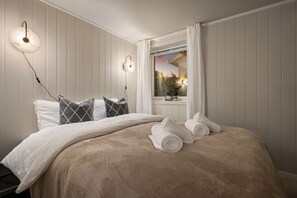 Un llit ben fet amb una manta i coixins beix, una taula de nit amb una làmpada i una finestra amb cortines.