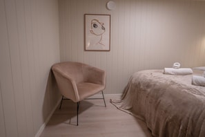Ein Zimmer mit einem einzelnen Sessel, einem Bett mit Decke und einem gerahmten Bild an der Wand.