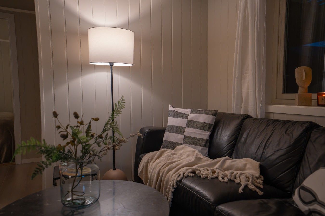 Un salon avec un canapé en cuir, une table en verre ornée d’une plante, un lampadaire et une fenêtre avec des rideaux.