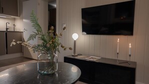A modern living room with a round table, a vase with dried flowers, a flat-screen TV mounted on the wall, and two candles on a side table. - Sydpol Designer Home Deluxe Apartment (Tromsø)