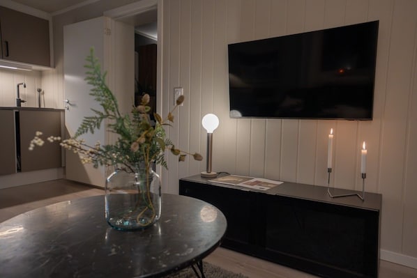 Ein modernes Wohnzimmer mit einem runden Tisch, einer Vase mit Trockenblumen, einem flachbildfernseher an der Wand und zwei Kerzen auf einem Beistelltisch.
