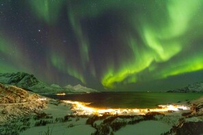 A snowy landscape with a lit area in the foreground, likely a hotel or resort, and a view of the night sky with the Northern Lights. - Modern 1BR Apartment – Cosy South Tromsøya Stay (Tromsø)