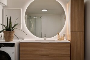 A modern bathroom with a round mirror, wooden cabinetry, and a plant on the counter.