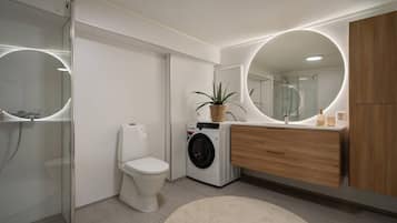 A modern bathroom with a glass shower enclosure, a toilet, a washing machine, and a wooden vanity with a mirror.