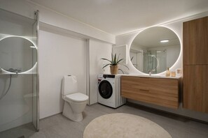 A modern bathroom with a glass shower enclosure, a toilet, a washing machine, and a wooden vanity with a mirror. - Sydpol Designer Home Classic Apartment (Tromsø)