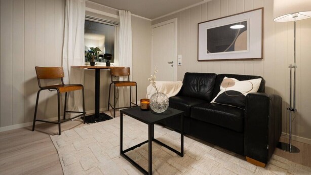 Un salon moderne avec un canapé en cuir noir, deux fauteuils en bois, une petite table basse noire, un lampadaire et un tableau abstrait encadré accroché au mur.