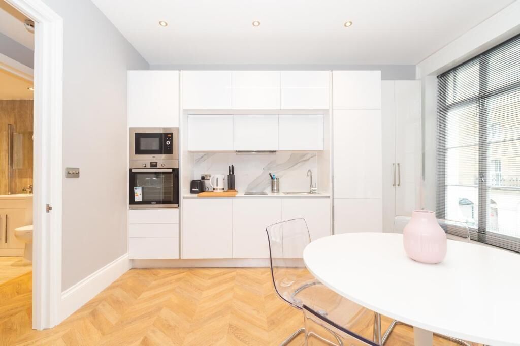 Een moderne keuken met witte kastjes, een magnetron en een koffiezetapparaat. Er staat een eettafel met stoelen en een roze vaas.