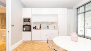 Een moderne keuken met witte kastjes, een magnetron en een koffiezetapparaat. Er staat een eettafel met stoelen en een roze vaas.