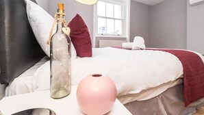 Un dormitorio con una cama, una mesita de noche con una botella y un jarrón rosa, una ventana y una almohada blanca.