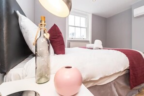 Un dormitorio con una cama, una mesita de noche con una botella y un jarrón rosa, una ventana y una almohada blanca.