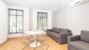 A modern living room with a round table, two grey sofas, and a large window with blinds.