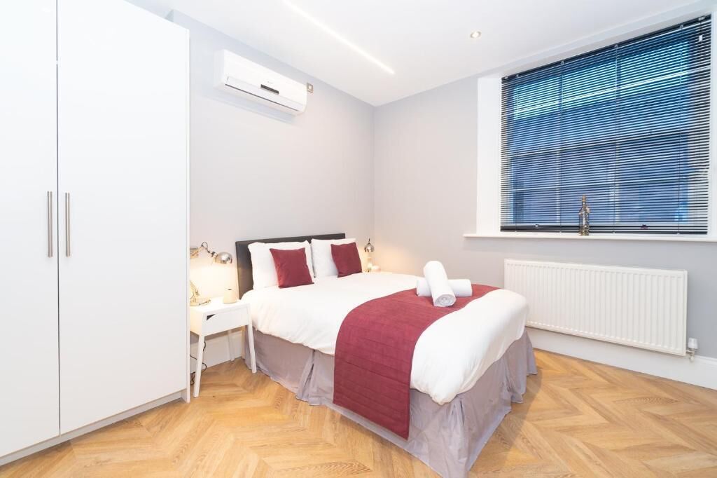 A hotel room with a bed, bedside table, lamp, and window with blinds.