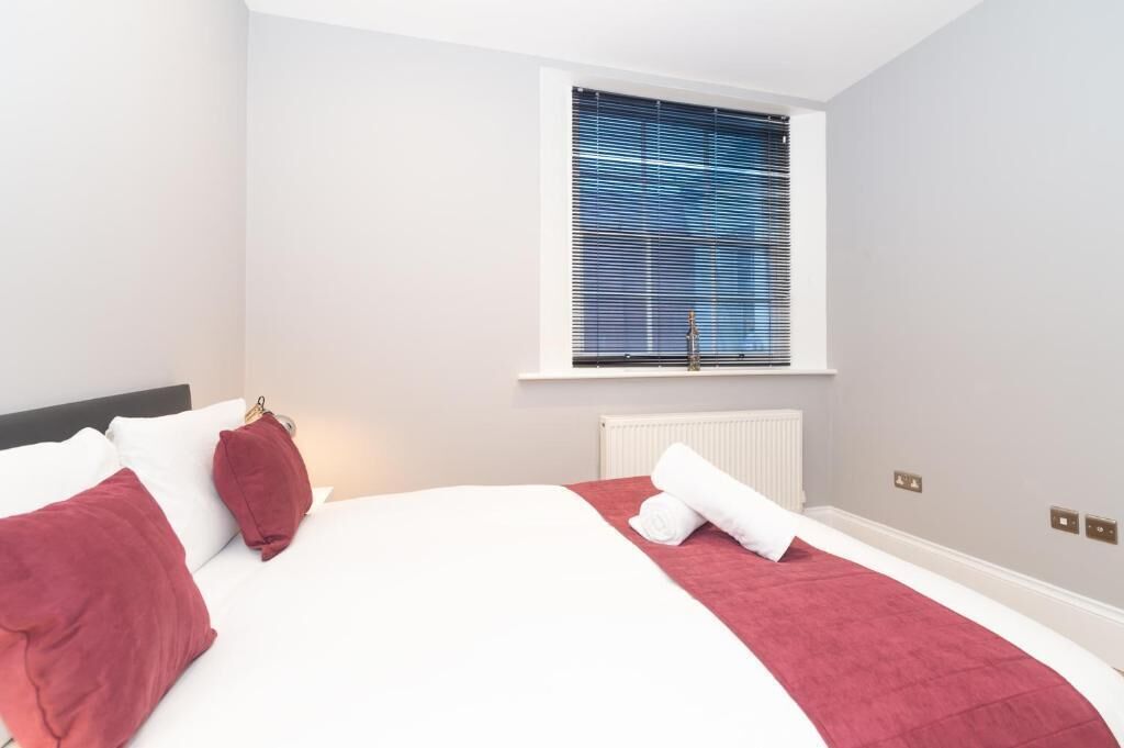 A hotel room with a bed, a window with blinds, a radiator, and a wall-mounted light switch.