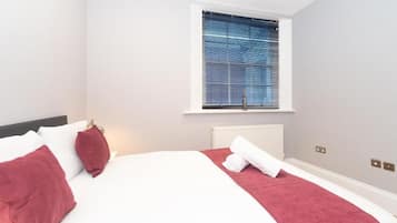 A hotel room with a bed, a window with blinds, a radiator, and a wall-mounted light switch.