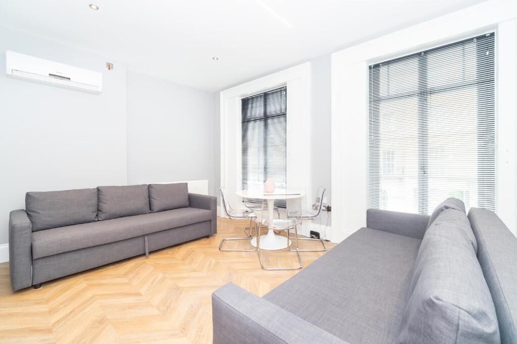 Una sala moderna con un sofá gris, una mesa de comedor con sillas y ventanas amplias con persianas.