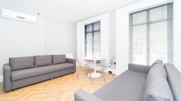 Una sala moderna con un sofá gris, una mesa de comedor con sillas y ventanas amplias con persianas.