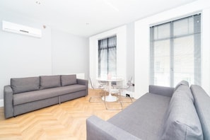 Una sala moderna con un sofá gris, una mesa de comedor con sillas y ventanas amplias con persianas.